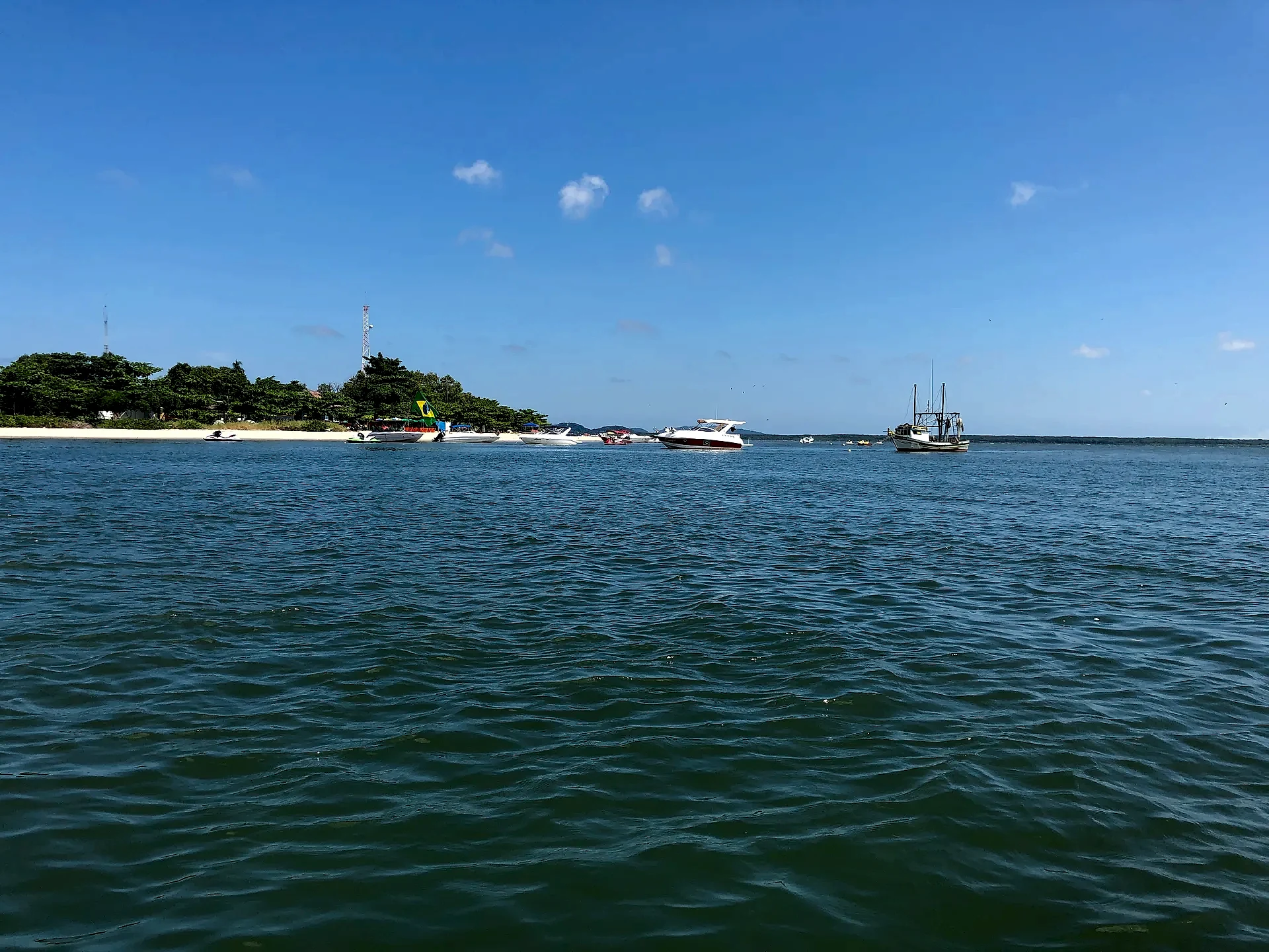 Barcos de passeio e uma embarcação de pesca navegam diante de uma faixa de praia com mata verde ao fundo. O mar calmo e o céu azul quase sem nuvens recebem luz clara de dia ensolarado.