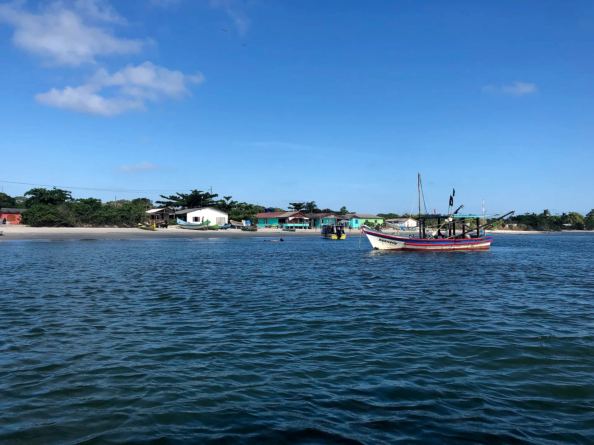 Barco de pesca ancorado em frente a uma pequena vila à beira da praia, com outras embarcações na areia e vegetação ao fundo. O mar levemente ondulado e o céu azul com poucas nuvens recebem luz clara de dia ensolarado.