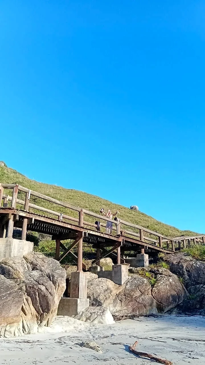 Passarela de madeira sobre formações rochosas junto à praia, com uma pessoa apreciando a vista e vegetação baixa no morro ao fundo. O dia ensolarado tem céu intensamente azul e luz clara sobre a areia e as pedras.