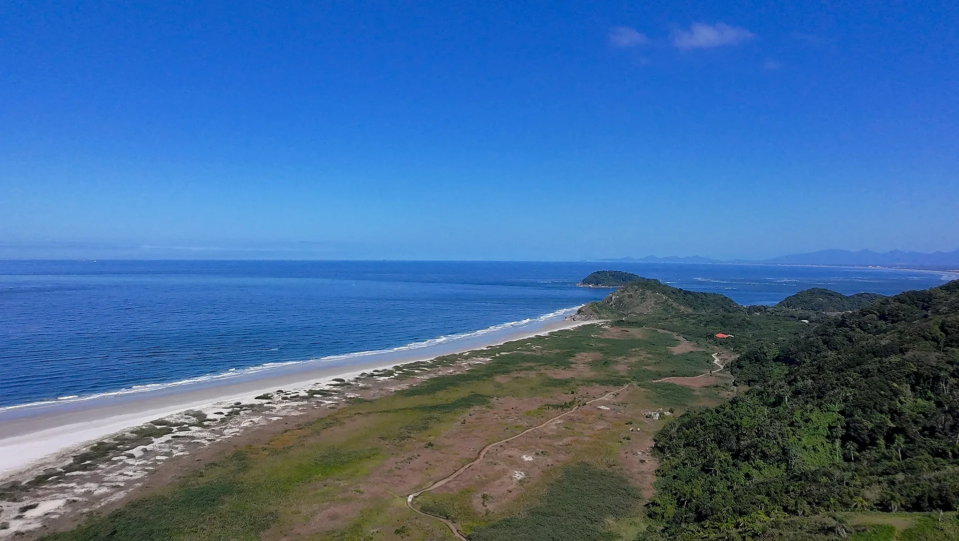 Vista aérea de praia de areia clara junto ao mar, com trilha atravessando área de vegetação baixa e mata densa nas encostas. A luz diurna intensa realça os tons azuis da água e o verde da paisagem costeira.