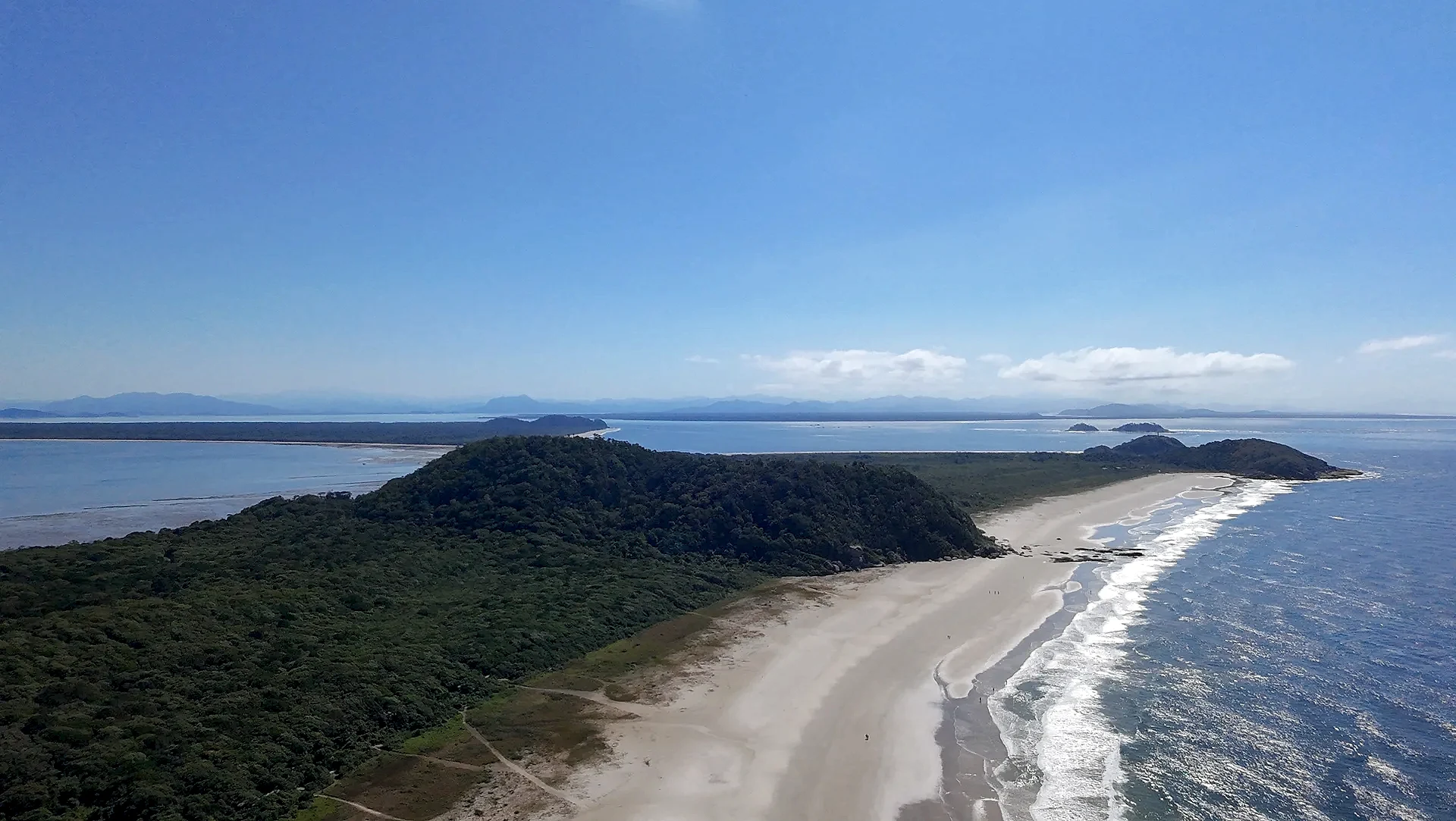 Vista aérea de uma praia extensa, com faixa de areia clara, mar azul e vegetação rasteira que avança até a mata. Uma trilha corta o terreno em direção à costa, sob luz solar intensa e céu limpo.