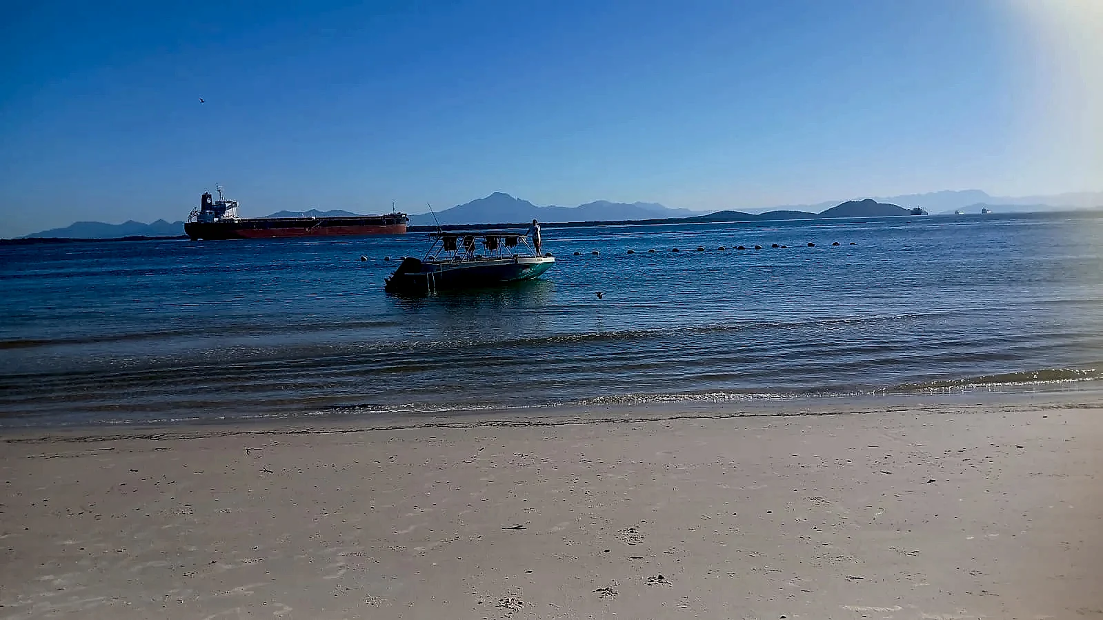 Praia de areia clara e mar calmo, com um pequeno barco ancorado próximo à faixa de arrebentação e uma pessoa a bordo. Ao fundo, navio cargueiro e montanhas no horizonte sob luz suave de céu azul.