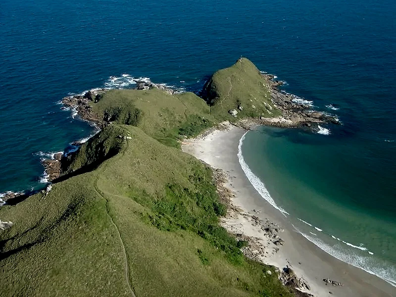Vista aérea de uma pequena praia entre morros verdes e costões rochosos, cercada pelo mar de tons azulados. A luz clara do dia destaca a trilha na vegetação, a faixa de areia e as ondas suaves na enseada.