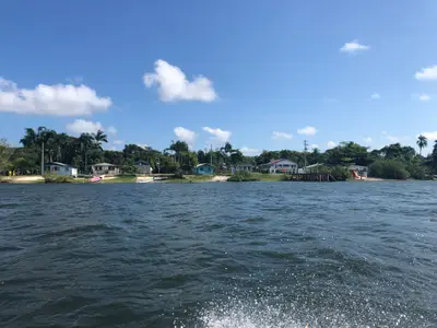 Vila à beira d’água, com casas simples, vegetação tropical e pequenas embarcações junto à margem. A cena é iluminada por sol forte sob céu azul com nuvens esparsas.