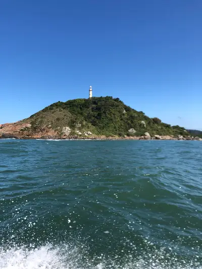 Farol sobre morro coberto pela mata, com costão rochoso à beira-mar visto a partir de um barco. A luz intensa do dia realça o céu azul, a água verde-azulada e a espuma das ondas.