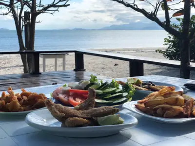 Pratos de frutos do mar, salada e batatas são servidos em mesa à beira da praia, com o mar e montanhas ao fundo. O ambiente sombreado por árvores recebe luz suave de dia parcialmente nublado.