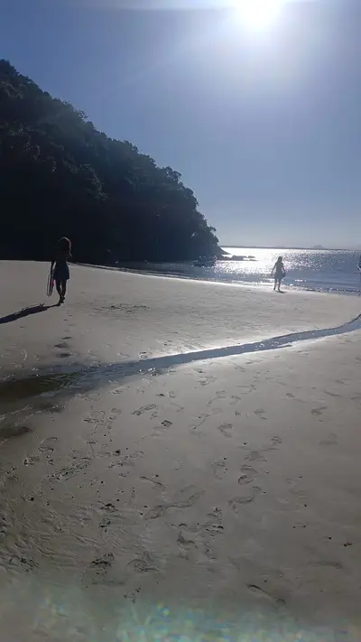 Praia de areia clara junto à mata, com duas pessoas caminhando perto da água e marcas de passos na faixa de areia. A luz forte do sol cria reflexos intensos no mar e sombras longas, em um ambiente tranquilo.