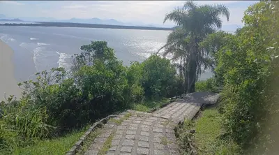 Trilha calçada desce pela mata costeira, com palmeiras e vegetação abundante em primeiro plano. Ao fundo, a praia e o mar refletem a luz suave do dia, com montanhas no horizonte.