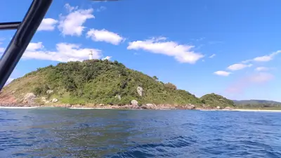 Farol no alto de um morro coberto por mata, visto do mar a partir de um barco, com costões rochosos e pequena faixa de praia ao fundo. A cena tem luz solar intensa, céu azul e nuvens esparsas.