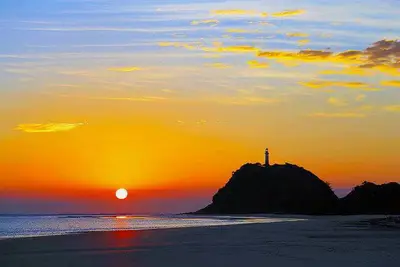 Pôr do sol sobre a praia, com o mar calmo e a silhueta de um morro coroado por um farol ao fundo. A luz dourada e alaranjada colore o céu e se reflete na areia úmida e na água.