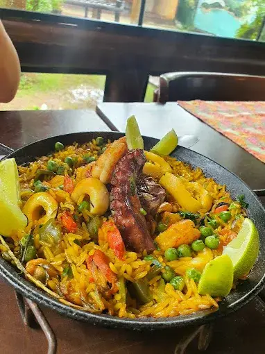 Prato de arroz com frutos do mar, legumes e gomos de limão servido em uma mesa de restaurante. O ambiente interno é acolhedor, com luz natural entrando pelas janelas e vegetação ao fundo.
