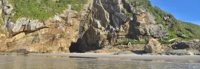 Praia de faixa estreita ao pé de um costão rochoso coberto por mata, com escadaria íngreme cravada nas pedras e pequena estrutura de madeira à direita. A luz solar direta destaca os tons dourados das rochas e o mar calmo em primeiro plano.