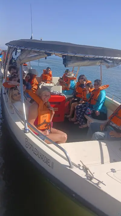 Grupo de pessoas com coletes salva-vidas em barco de passeio sobre águas calmas, sob cobertura de lona. A cena é iluminada por sol forte, com céu azul e clima de passeio marítimo.